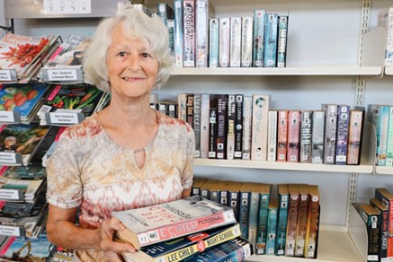 Librarian Sherril is holding a stack of three books, with a library bookshelf in the background