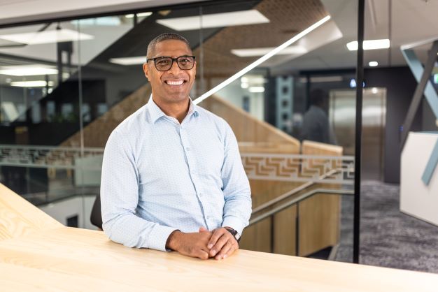 Senior leader stands behind a wooden bench in our National Office with glass panels decorated with the Poutama staircase design in the background